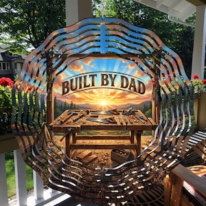 May include: A metal wind spinner with a layered, wavy design. The center features a detailed image of a woodworking bench with tools and the text "BUILT BY DAD" against a sunset backdrop. The spinner hangs on a porch.