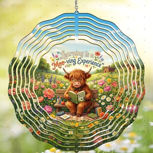 May include: A layered, circular ornament featuring a cartoon Highland cow reading a book on a bench in a flower-filled meadow. The text "Learning is a Moo-ving Experience" is displayed above. The ornament has a blue sky design at the top and is suitable for hanging.
