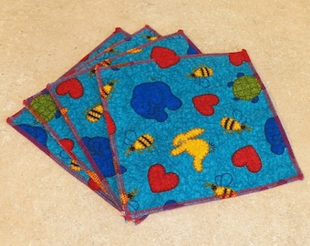 Puede incluir: Un conjunto de cuatro cuadrados de tela azul con un patrón rojo, amarillo y verde. Los cuadrados presentan corazones, abejas, conejos y tortugas.