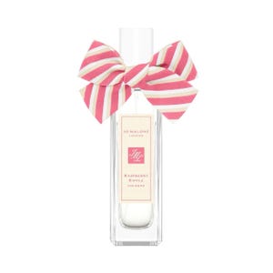 Pode incluir: Um frasco de perfume retangular de vidro transparente com um rótulo branco contendo o texto Jo Malone London, JML e Raspberry Ripple Cologne. Um grande laço de tecido com listras diagonais rosa, brancas e bege está atado ao gargalo.