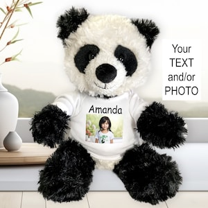 May include: A plush panda bear with black and white fur, wearing a white t-shirt with a photo and the name "Amanda". The bear has black paws and ears. The background includes a window and decorative vases.