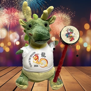 Puede incluir: Un dragón de peluche verde que lleva una camiseta blanca con el texto "Year of the Dragon 2024" y un gráfico de dragón chino rojo. El dragón sostiene un tambor chino rojo y negro con un palo.