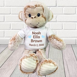 May include: A plush monkey toy with a white t-shirt. The shirt has the name "Noah Ellis Brown" and the date "March 2, 2026" printed on it, with blue heart accents. The monkey is light brown and beige.