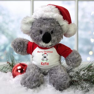 May include: A gray koala plush toy wearing a red and white Santa hat and a white shirt with the text "Merry Christmas Katie". The toy is sitting in a snowy scene with a red Christmas ornament.