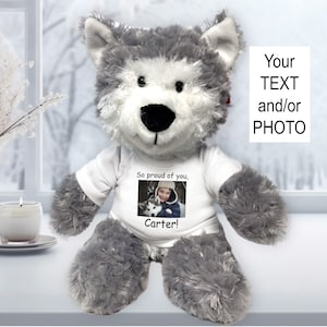 May include: A gray and white plush wolf wearing a white t-shirt with a photo of a child and a dog and the text "So proud of you, Carter!"