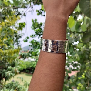 May include: A silver-colored cuff bracelet with a hammered texture. The wide band encircles the wrist, reflecting light. The background is a blurred view of green foliage and a bright sky.