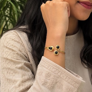 May include: A gold-toned open cuff bracelet featuring three smooth, round black stones. The metal band curves into a spiral design at the ends, with one stone set at the single terminal and two stones set at the double-ended terminal of the cuff.