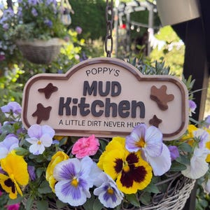 May include: A rectangular beige sign with rounded corners and a dark brown border, featuring the text "POPPY'S MUD KITCHEN" and "A LITTLE DIRT NEVER HURT" in dark brown lettering, accented by small brown mud-splatter shapes on the sides.