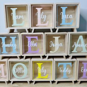 May include: A set of 10 wooden boxes with clear glass fronts. Each box has a different name written on it in white letters: Mia, Ollie, Lily, Teddy, Esmae, Huxley, Ella, Jorja, Archie, Liam, Lily, Luca.