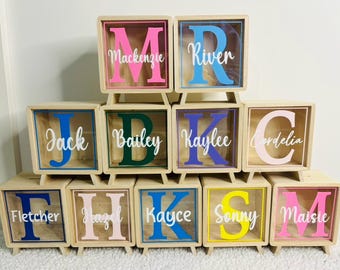 May include: A collection of square wooden shadow boxes with clear fronts, each featuring a large colourful letter and a white script name. The boxes are arranged in a pyramid stack, displaying various names including Mackenzie, River, Jack, Bailey, Kaylee, Cordelia, Fletcher, Hazel, Kayce, Sonny, and Maisie.