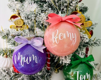 May include: Three round, glittery Christmas baubles in purple, peach, and green. Each bauble features a white script name: Mum, Remy, and Tom. Each is topped with a matching satin ribbon bow and a silver cap, hanging from a flocked tree.