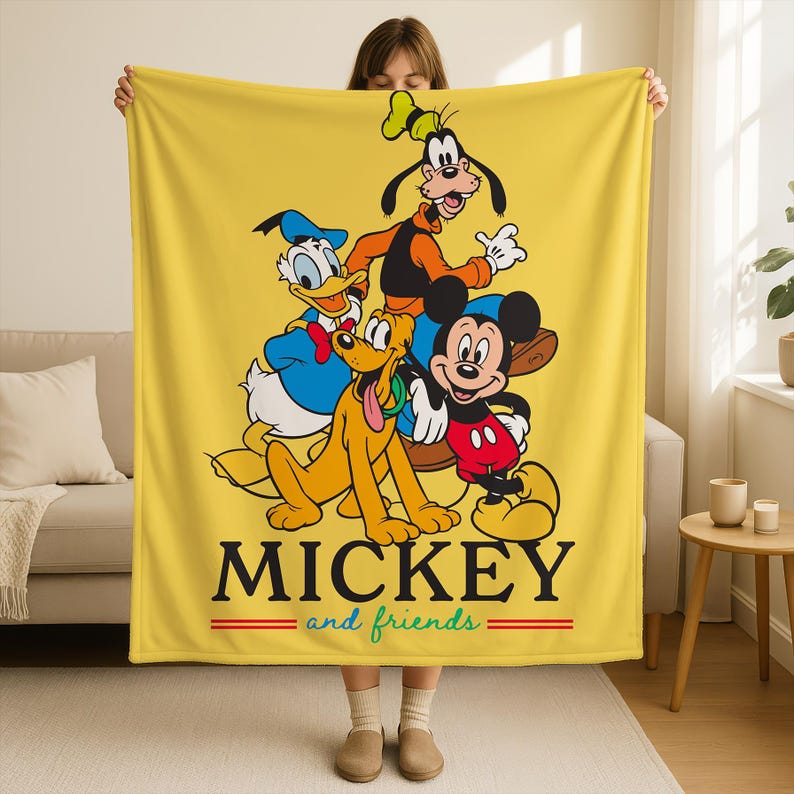 May include: Yellow blanket featuring Mickey Mouse, Donald Duck, Goofy, and Pluto. The blanket has the words "MICKEY and friends" in black with red and green accents. The blanket is held up by a person.