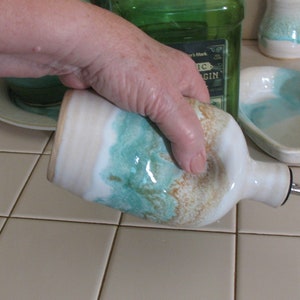 May include: A ceramic oil bottle with a white base, teal and brown accents, and a metal spout. The bottle is held by a hand, suggesting its use. Other ceramic kitchenware is in the background.