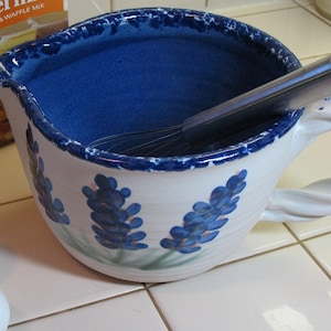 May include: A white ceramic mixing bowl with a blue rim and blue floral design. The bowl has a handle and a whisk inside. The bowl is sitting on a white tiled surface.