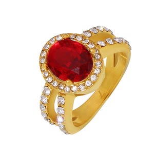 May include: A gold-toned ring featuring a large, oval-cut red gemstone at the center, surrounded by a halo of smaller, clear gemstones. Additional clear gemstones are set along the ring's band, creating a sparkling effect. The ring has a classic, elegant design.