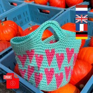Op de afbeelding: Een turquoise gehaakte draagtas met roze hartjes. De tas heeft een stevige handgreep en wordt getoond in een blauwe krat met verschillende oranje pompoenen. Een PDF-pictogram is zichtbaar linksonder.