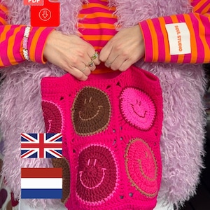 Op de afbeelding: Een felroze gehaakte tas opgebouwd uit samengevoegde vierkante motieven, elk met een cirkelvormig smiley-gezicht in contrasterende tinten bruin, donkerroze en lichtroze. De tas heeft een getextureerd, handgemaakt uiterlijk met zichtbare steken.