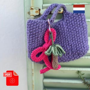 Op de afbeelding: Een kleine, handgehaakte paarse draagtas met een gevlochten handvat. Een roze en groene gehaakte bedel is met een zilverkleurige sluiting aan de tas bevestigd. De tas hangt aan een verweerde, lichtgroene luik.