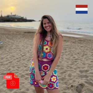 Op de afbeelding: Een kleurrijke gehaakte tweedelige outfit met een bloemenpatroon. De top en rok zijn in verschillende kleuren, waaronder paars, geel, groen en blauw. De outfit wordt gedragen op een strand met een zonsondergang op de achtergrond.