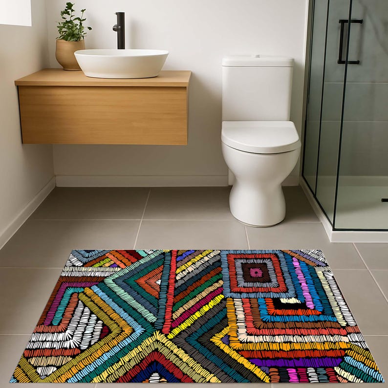 May include: A rectangular bathroom rug with a vibrant, geometric design. The rug showcases a patchwork of embroidered patterns in various colours, including blue, green, red, yellow, and black. The rug is placed on a grey tiled floor.