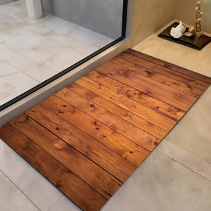 K&ouml;nnte beinhalten: Eine rechteckige Fu&szlig;matte mit Holzplanken-Design. Die Matte zeigt ein Muster aus parallelen, dunkelbraunen Holzplanken mit sichtbaren Knoten und Maserungen. Die Matte liegt auf einem hell gefliesten Boden in der N&auml;he einer Glast&uuml;r.