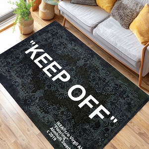 以下が含まれることがあります： 大きな白い文字で「KEEP OFF」と書かれた、花柄の黒い長方形のラグ。ラグには「IKEA c/o Virgil Abloh Flooring Almhult Swed c 2019」というテキストも含まれています。
