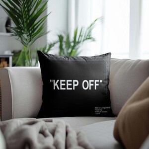 May include: Black throw pillow with white text that reads "KEEP OFF" on a gray couch. The pillow has a black and white patterned border.