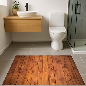 K&ouml;nnte beinhalten: Ein rechteckiger Badezimmerteppich mit Holzplanken-Design. Der Teppich zeigt vertikale Planken in verschiedenen Braunt&ouml;nen, wodurch ein realistisches Holzmaserungs-Aussehen entsteht. Der Teppich liegt auf einem grau gefliesten Boden in einem Badezimmer.