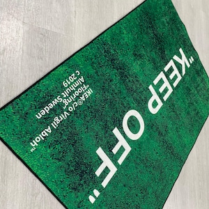 Puede incluir: Una alfombra verde con el texto "KEEP OFF" en letras blancas. La alfombra est&aacute; dise&ntilde;ada para parecer c&eacute;sped y tiene el texto "IKEA&reg;c/o Virgil Abloh&trade; Almhult Sweden "Flooring" c 2019" impreso en la esquina inferior derecha.