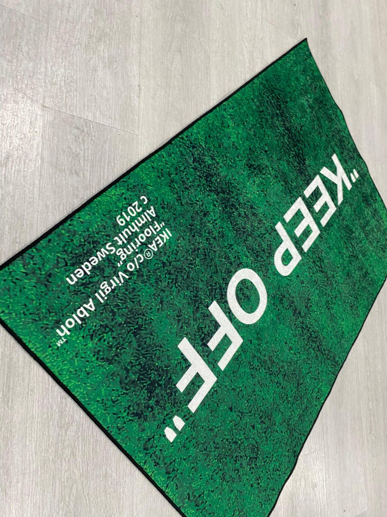 Puede incluir: Una alfombra verde con el texto "KEEP OFF" en letras blancas. La alfombra est&aacute; dise&ntilde;ada para parecer c&eacute;sped y tiene el texto "IKEA&reg;c/o Virgil Abloh&trade; Almhult Sweden "Flooring" c 2019" impreso en la esquina inferior derecha.