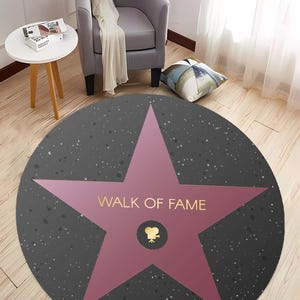May include: A round, gray and pink rug with a large pink star in the center. The star has the words "Walk of Fame" written on it in gold lettering. The star also has a small gold film camera icon in the center.