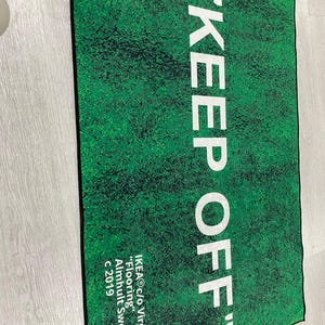 Puede incluir: Una alfombra de bienvenida verde y blanca con el texto "KEEP OFF" en blanco. La alfombra tambi&eacute;n incluye el texto "IKEA&reg;c/o Virgil Abloh&trade;" "Flooring" Almhult Sweden c 2019.