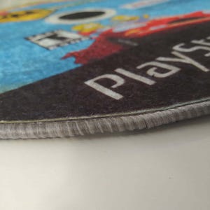 May include: Close-up of the edge of a round rug with a gray border. The rug has a printed image of a video game disc with the word "Playstation" printed on it.