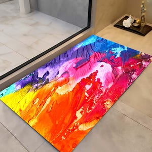May include: A rectangular floor mat with a vibrant abstract design. The mat features a blend of colours, including yellow, orange, red, pink, purple, and blue, creating a textured, artistic appearance. The mat is placed on a tiled floor.