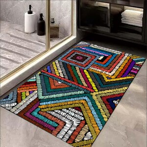 May include: A rectangular bath mat with a colourful geometric design. The mat features a patchwork of squares and angled lines in shades of red, orange, yellow, green, blue, purple, and white. The design is reminiscent of embroidery.