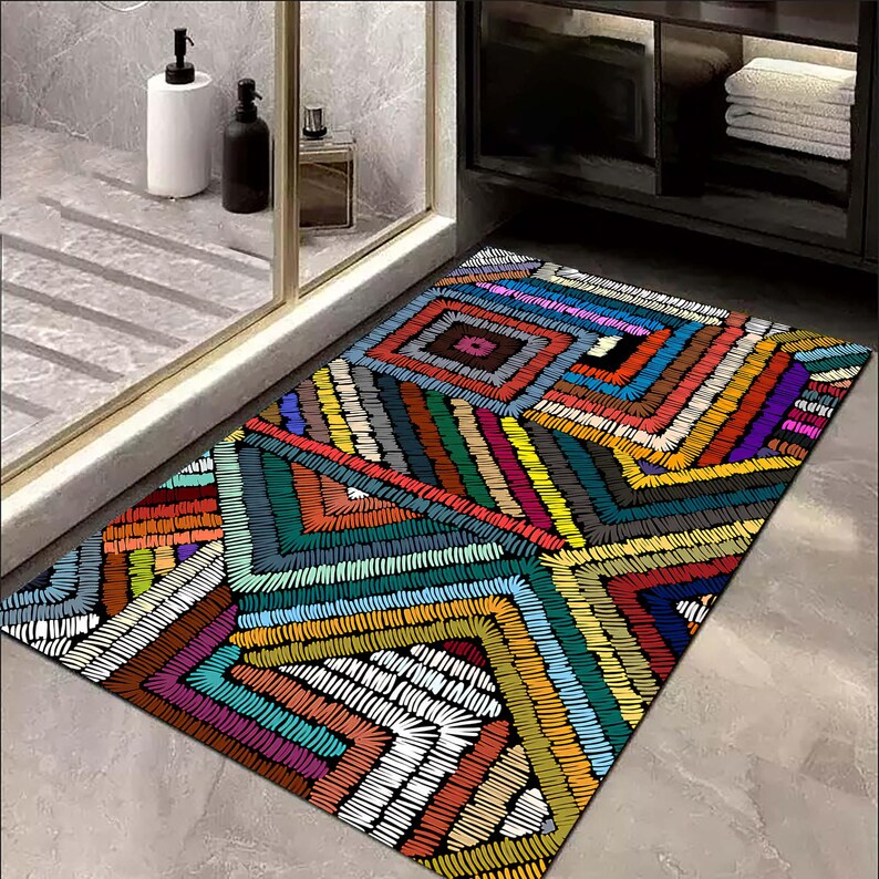May include: A rectangular bath mat with a colourful geometric design. The mat features a patchwork of squares and angled lines in shades of red, orange, yellow, green, blue, purple, and white. The design is reminiscent of embroidery.