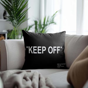 May include: Black throw pillow with white text that reads "KEEP OFF". The pillow is on a light gray couch with a brown blanket.