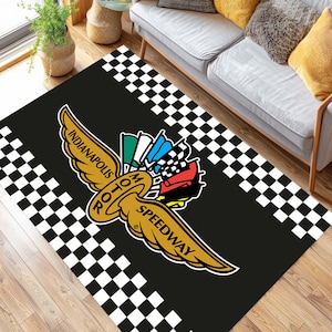 May include: Black rectangular rug featuring the Indianapolis Motor Speedway logo in gold, red, green, and blue. The logo includes the words "Indianapolis Motor Speedway" and "Roto". The rug has a black background with a checkered pattern border in white and black.