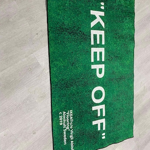 Puede incluir: Una alfombra verde con el texto "KEEP OFF" en letras blancas. La alfombra est&aacute; hecha por IKEA en colaboraci&oacute;n con Virgil Abloh y est&aacute; etiquetada como "Flooring" con el a&ntilde;o 2019.