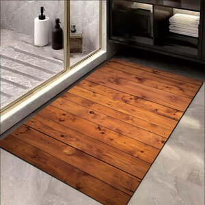 K&ouml;nnte beinhalten: Eine rechteckige Badematte mit Holzplanken-Design. Die Matte ist haupts&auml;chlich braun mit helleren Holzmaserungsdetails. Die Matte liegt auf einem grau gefliesten Boden in der N&auml;he einer Dusche und eines Schranks.