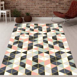 May include: A rectangular area rug with a geometric pattern of triangles in black, white, gray, pink, and light blue, outlined with gold lines. The rug is placed on a light-colored wooden floor.