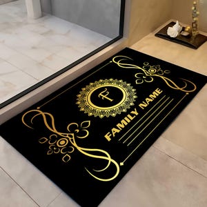 Peut inclure: Tapis de porte noir avec des détails dorés. Le tapis présente un emblème circulaire doré avec la lettre "F" et les mots "FAMILY NAME" en lettres dorées. Le design comprend des ornements.