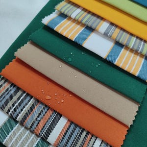 May include: A selection of fabric swatches in various colours and patterns, including stripes, plain colours, and textures. The swatches are stacked, with the top swatch being a white fabric with blue and yellow stripes.