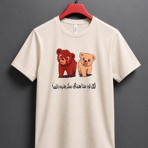 May include: A beige short-sleeved t-shirt featuring a graphic of two cartoon bears, one dark brown and one light tan, standing side by side. Below the bears is black Arabic text that reads: لكل لوز منا هناك سكر بقربه دائماً.