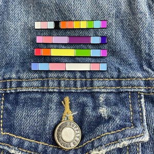 May include: Four enamel pins with different color palettes. Each pin is a horizontal bar with multiple colored squares. The pins are arranged vertically on a blue denim jacket.