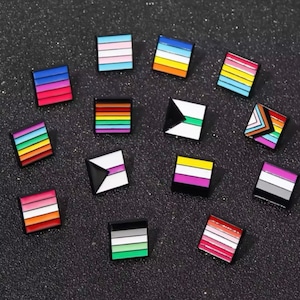 May include: A collection of square enamel pins with various LGBTQ+ pride flag designs. The pins feature vibrant colors and patterns, including rainbow stripes, transgender, and other pride flag representations. They are set against a dark, textured background.