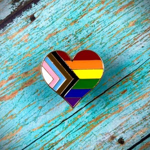 May include: A heart-shaped pin with a rainbow flag design, including the colors of the transgender, intersex, and pride flags.