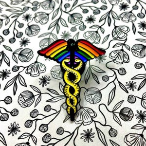 May include: A rainbow-colored medical caduceus pin with a yellow snake wrapped around the staff. The pin is on a white background with a black floral pattern.