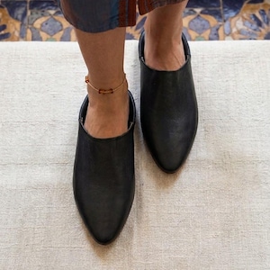 May include: A pair of black leather slip-on mules featuring a pointed toe design and a low-cut topline. The shoes have a smooth, matte finish and a simple, clean silhouette that covers the front of the foot while leaving the heel area exposed.