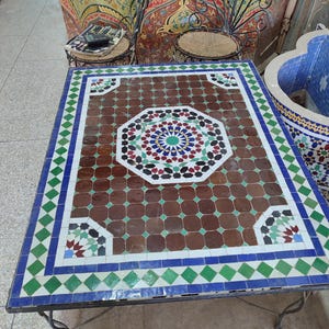 May include: A square table with a brown, green, and blue mosaic tile top. The table has a black metal frame and is sitting on a light-colored floor.